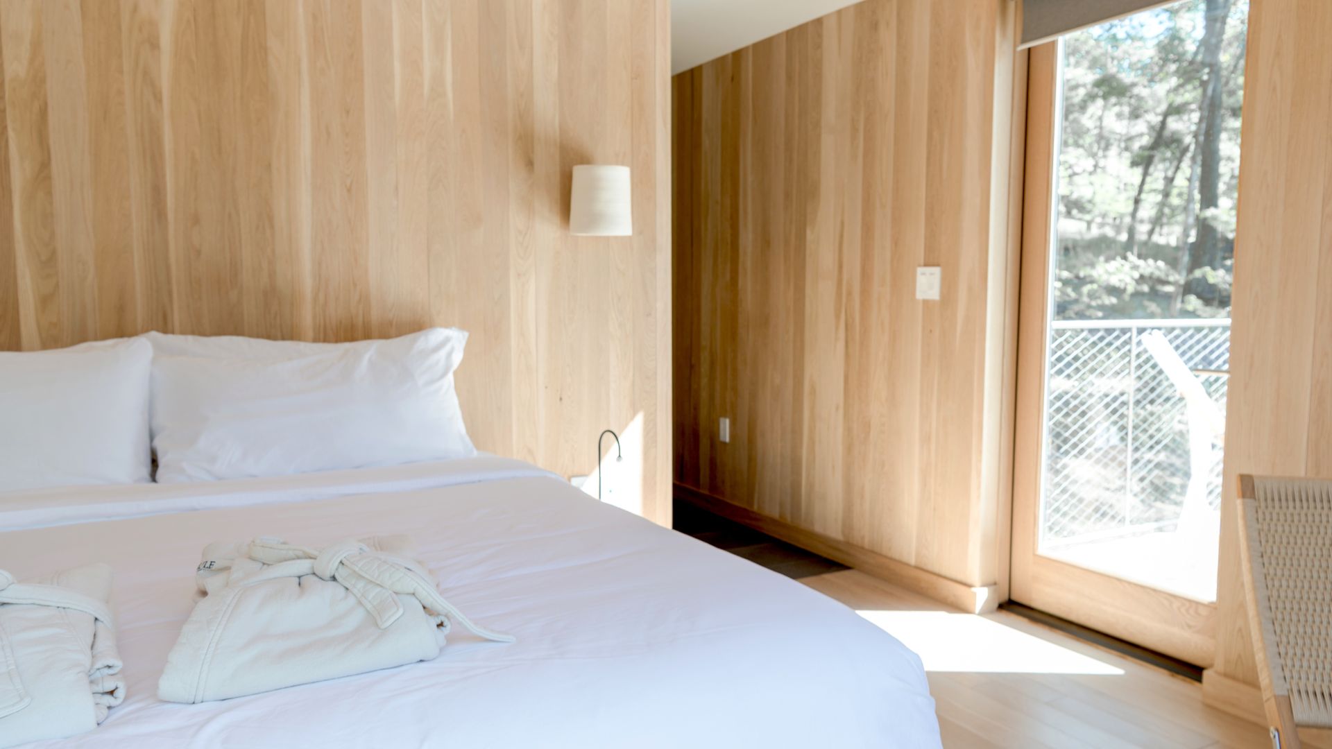 Minimalist wooden hotel room with white bed and large window overlooking trees